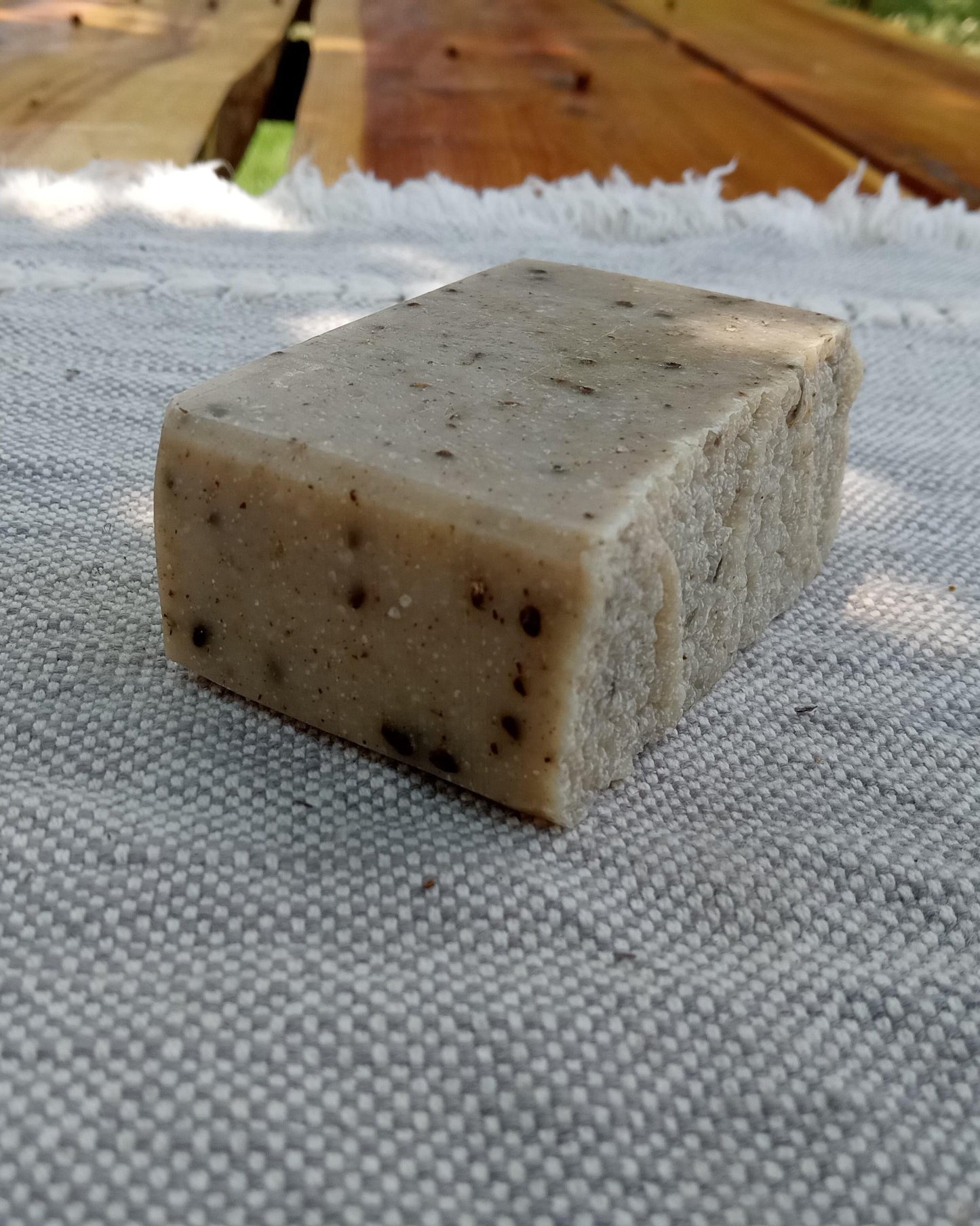 a beige bar of soap with flecks of dark brown in it, smooth edges and a rustic wrinkled top set on a woven cloth atop a rustic wooden table