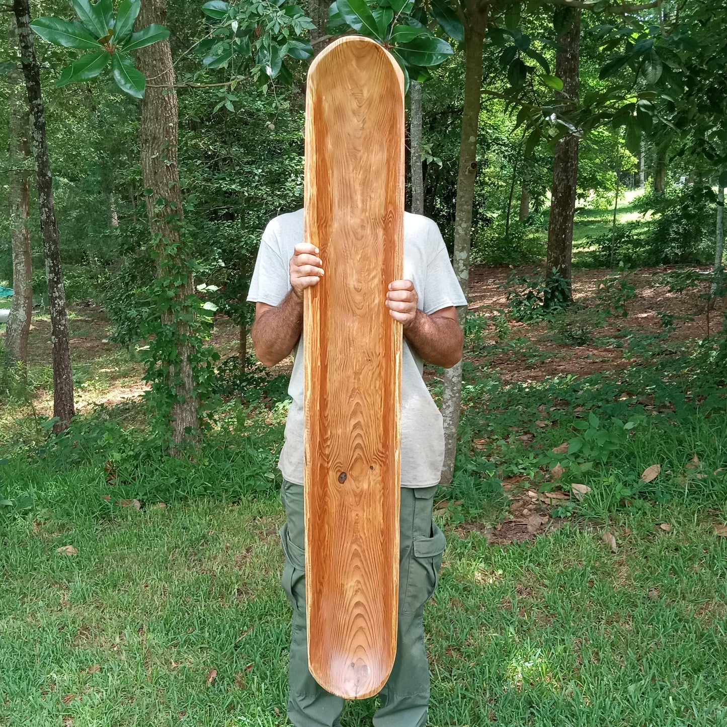 very long wooden trencher carved from heart pine with an antiqued orangey hue held by a man standing in the woods showing the piece to be about 5 feet long