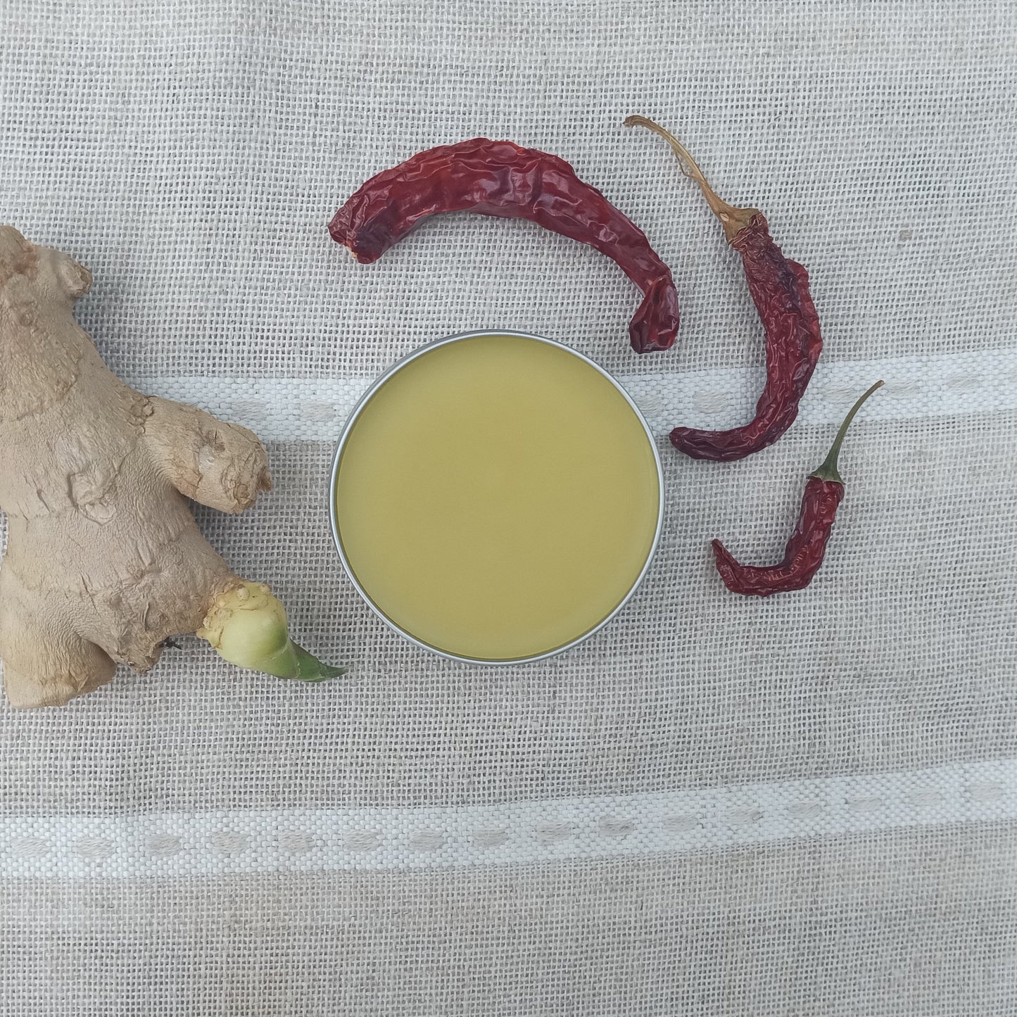 a tin of golden salve set on a woven linen table runner with a cayenne pepper and a ginger root set beside it