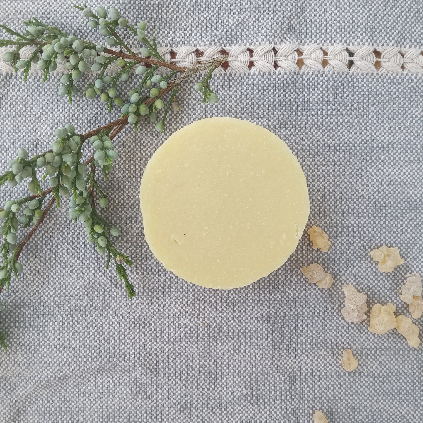 a creamy white round bar of soap set on pale blue woven fabric with a sprig of juniper berries and frankincense tears scattered around it