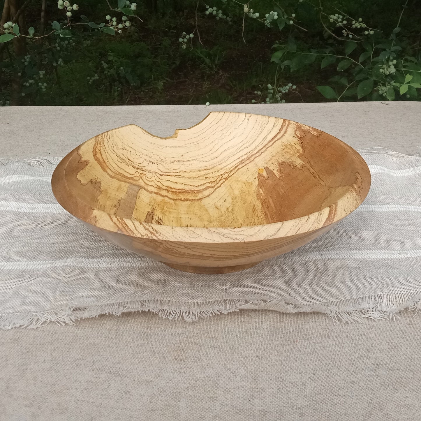 a highly figured shallow pecan wood bowl with cream and brown streaking details across the sides and a finely carved rim with a natural divot on one side set on a linen table runner with greenery in the background