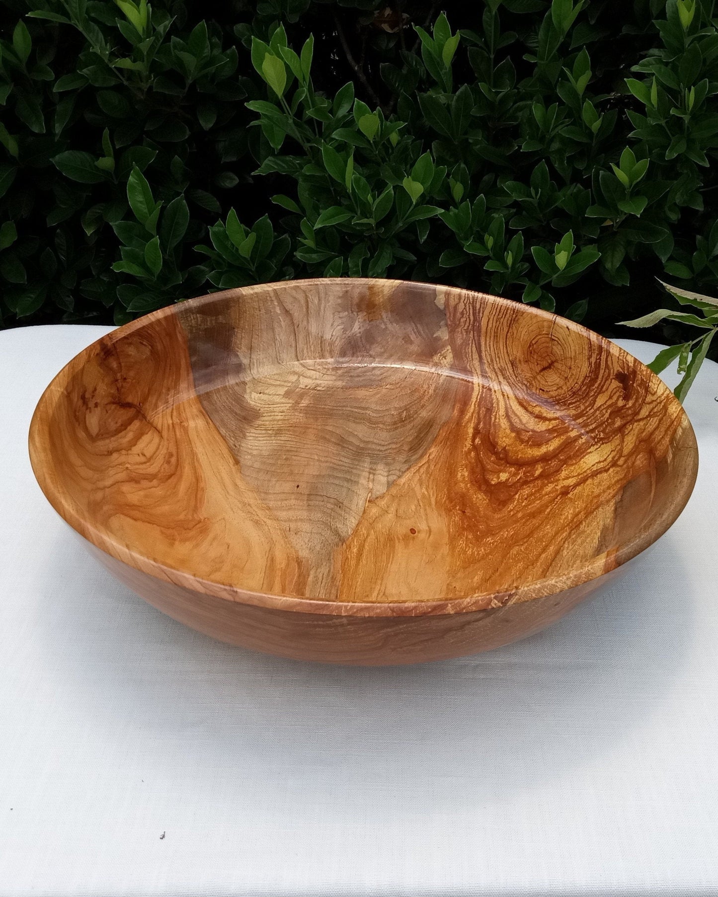 a huge highly figured shallow pecan wood bowl with stunning grain patterns that look like a volcanic eruption in hues of amber, burnt umber, burnt sienna and silvery gray set on a white tablecloth with greenery in the background