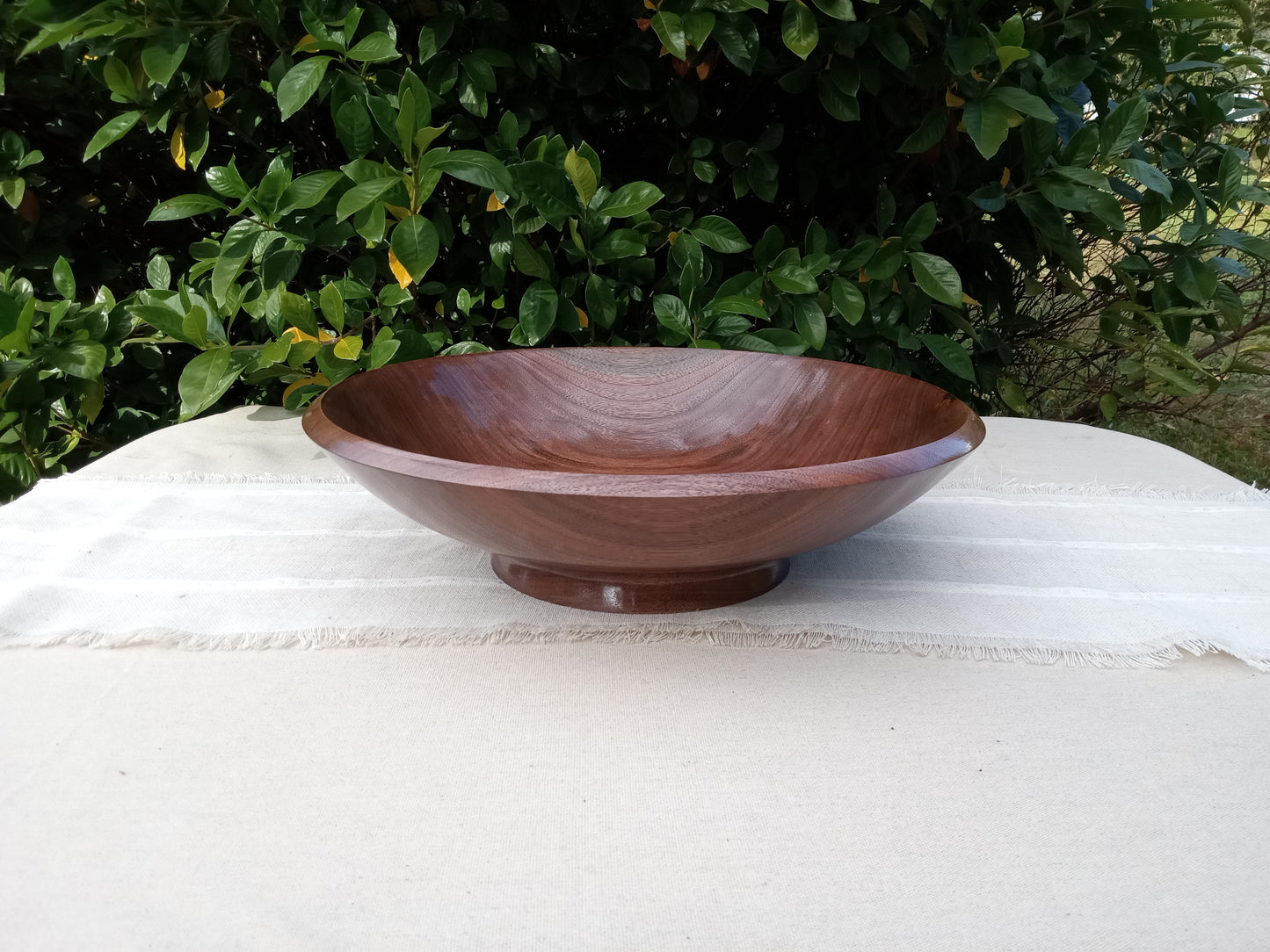 an elegant shallow black walnut wood pedestal bowl with a glossy finish and finely carved rim set on a natural linen table runner with greenery in the background