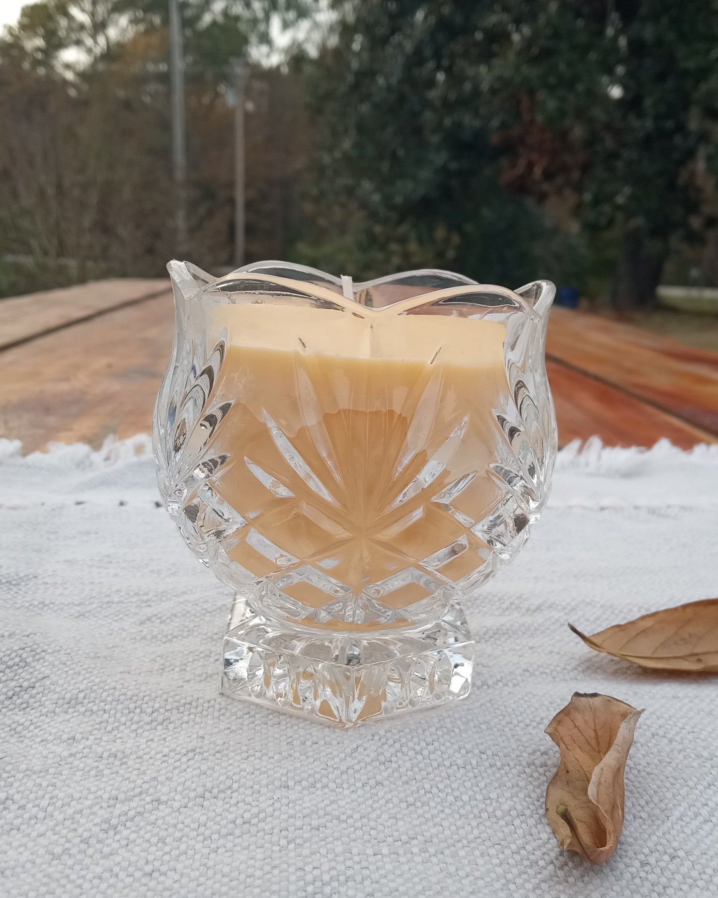 yellow beeswax candle in decorative crystal glass pedestal container with wavy edges set on a woven cloth atop a rustic wooden table with dried leaves scattered around