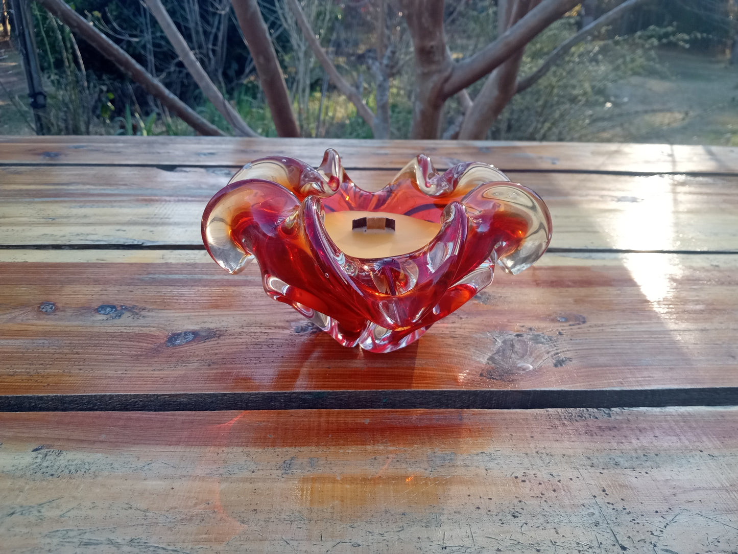 a yellow beeswax candle with a wooden wick in a red glass blown ashtray with petal like fluted edges set on a rustic wooden table