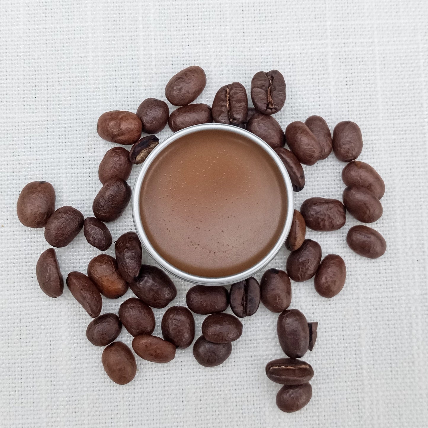 coffee colored salve in a silver tin set on white linen with a pile of coffee beans arranged around it