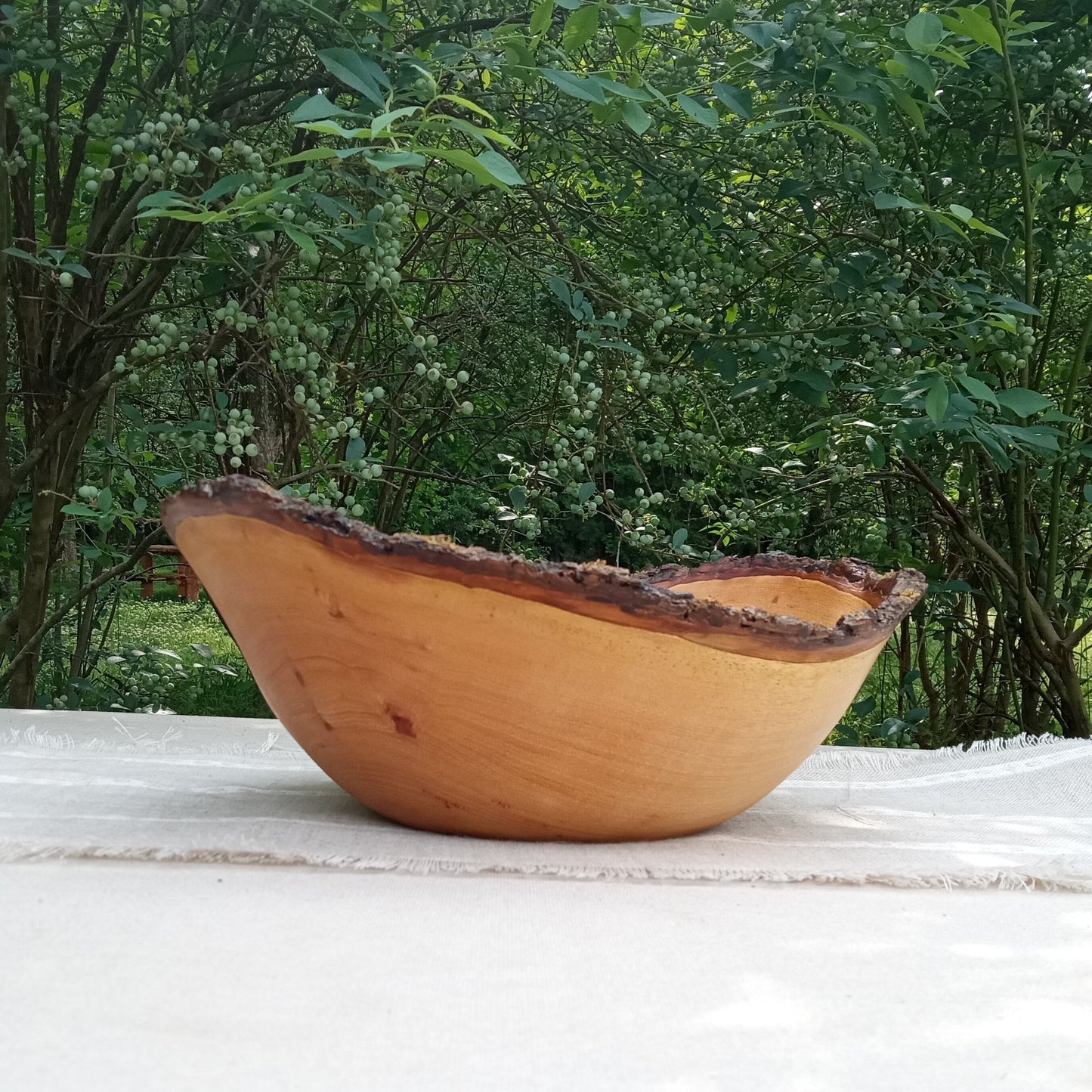 a light toned live edge wood bowl with dark jagged bark on the rim set on a white and beige linen table runner with blueberry bushes in the background