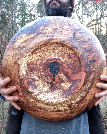 a massive oak burl carved into a huge round bowl with a slight footing carved into the bottom encircling the wild wood brand. burnt umber, creamy ochre, burnt sienna and peachy pink hues swirl in incredible spiraling patterns throughout the wood