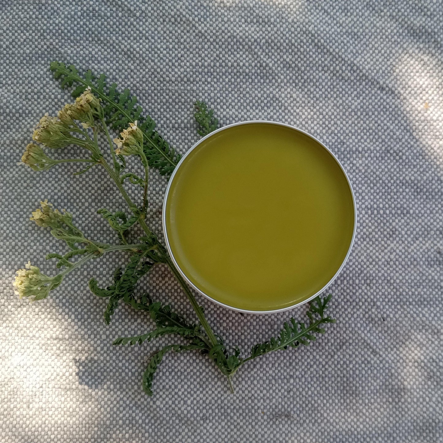 golden salve in a silver tin set on woven cotton with yarrow flowers and leaves