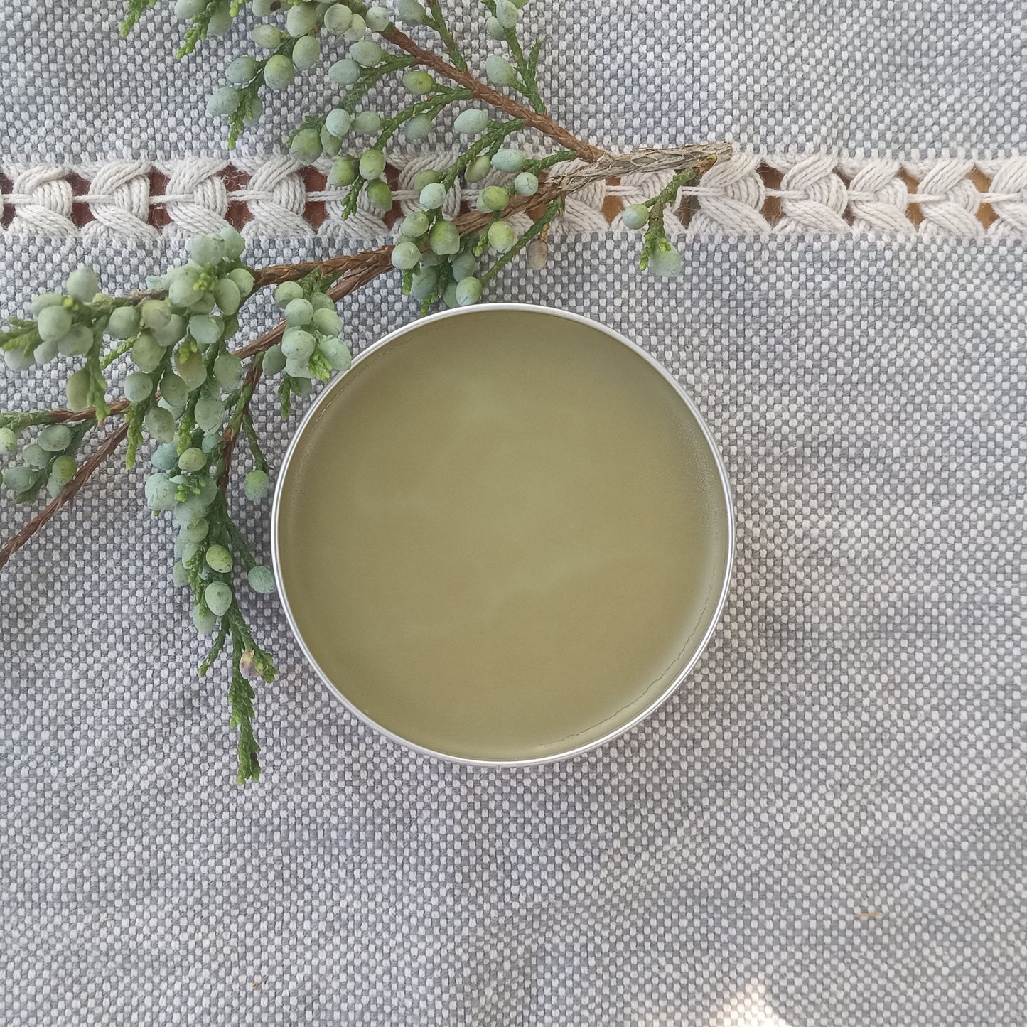 light beige salve in a silver tin on pale blue woven cotton with a sprig of juniper berries next to it