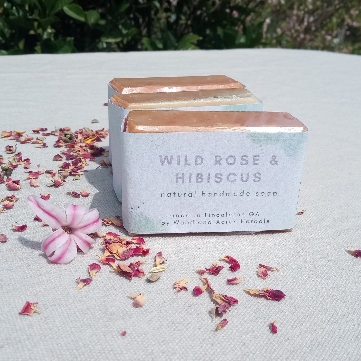 three rectangular pink soaps labeled "wild rose & hibiscus" with smooth shiny edges set atop a white tablecloth with pink rose petals and hyacinth buds scattered around