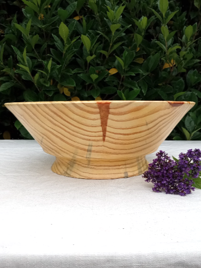 a large vase shaped light wood pedestal bowl with a V shaped streak of orange resin in the sidewall set on a white table cloth with purple heliotrope flowers next to it