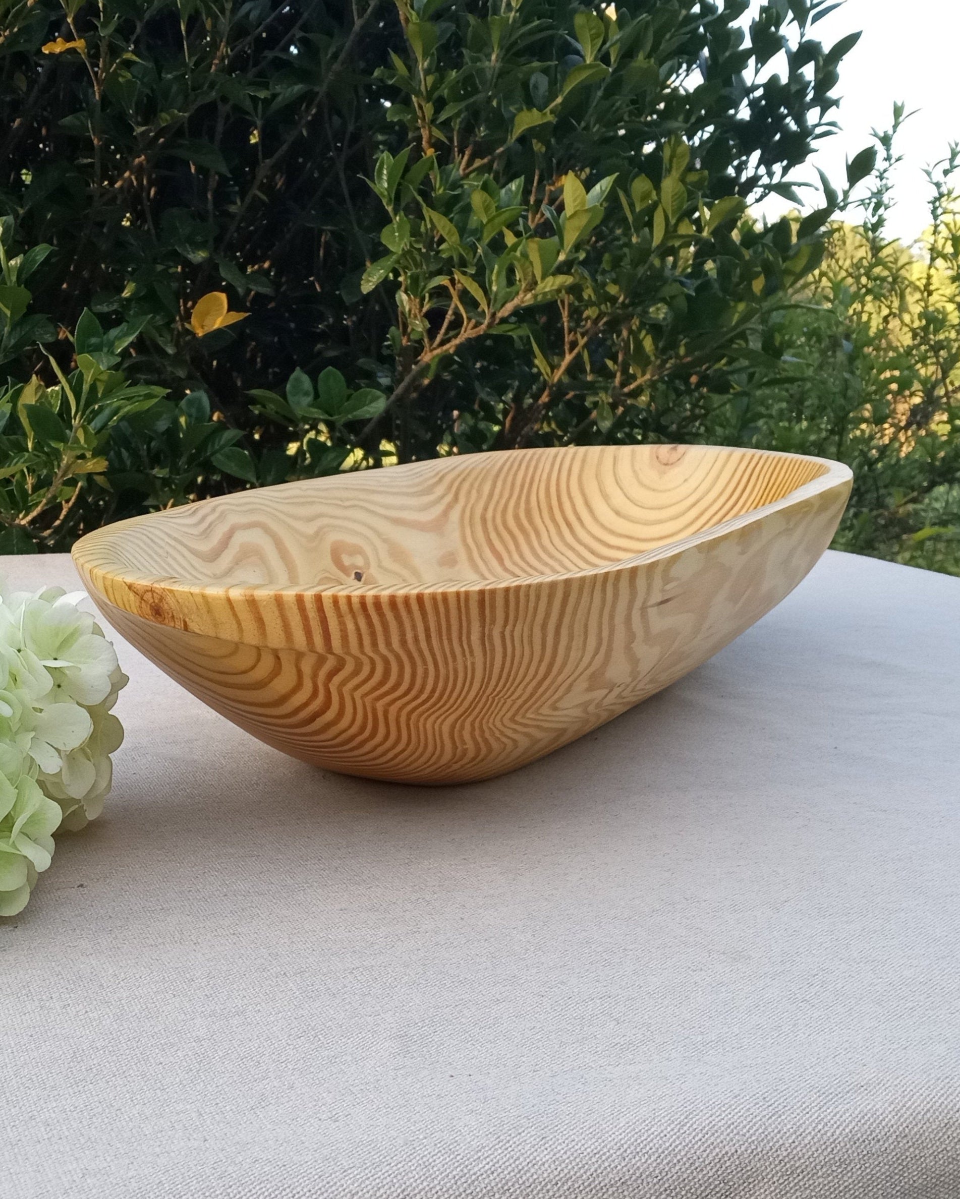 side view of an oblong plain pine wood dough bowl with handles and a deep sidewall set on a white table cloth next to a clump of hydrangea flowers