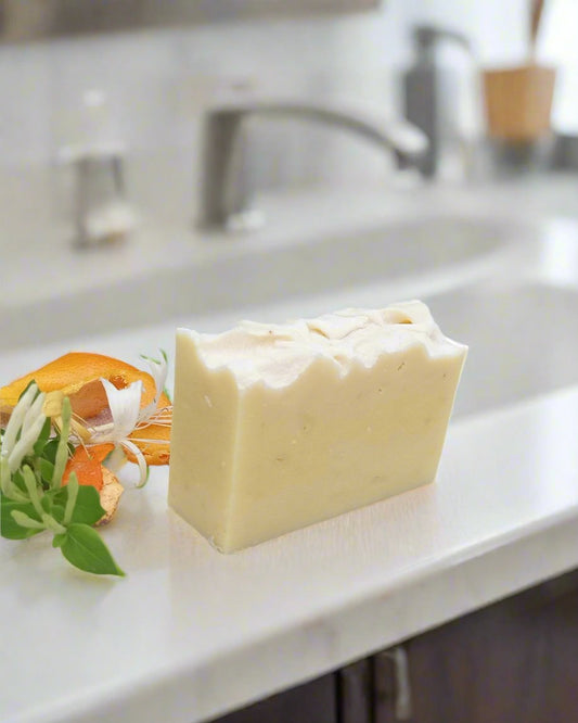 a creamy white rectangular bar of soap with a sculpted top set on a white bathroom counter with honeysuckle flowers and orange peels beside it