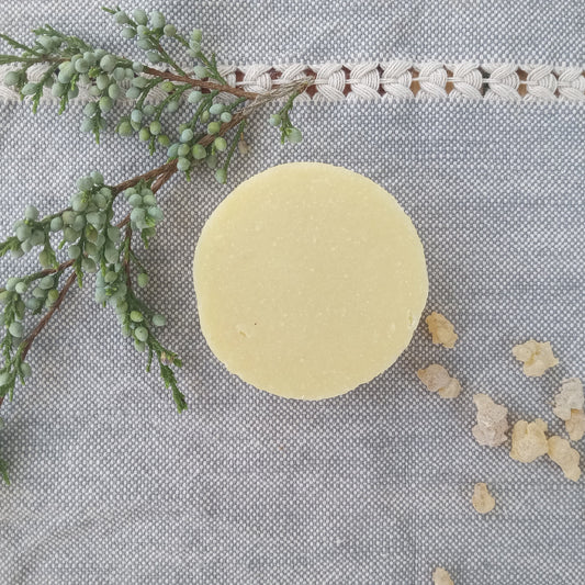 a creamy white round bar of soap set on pale blue woven fabric with a sprig of juniper berries and frankincense tears scattered around it