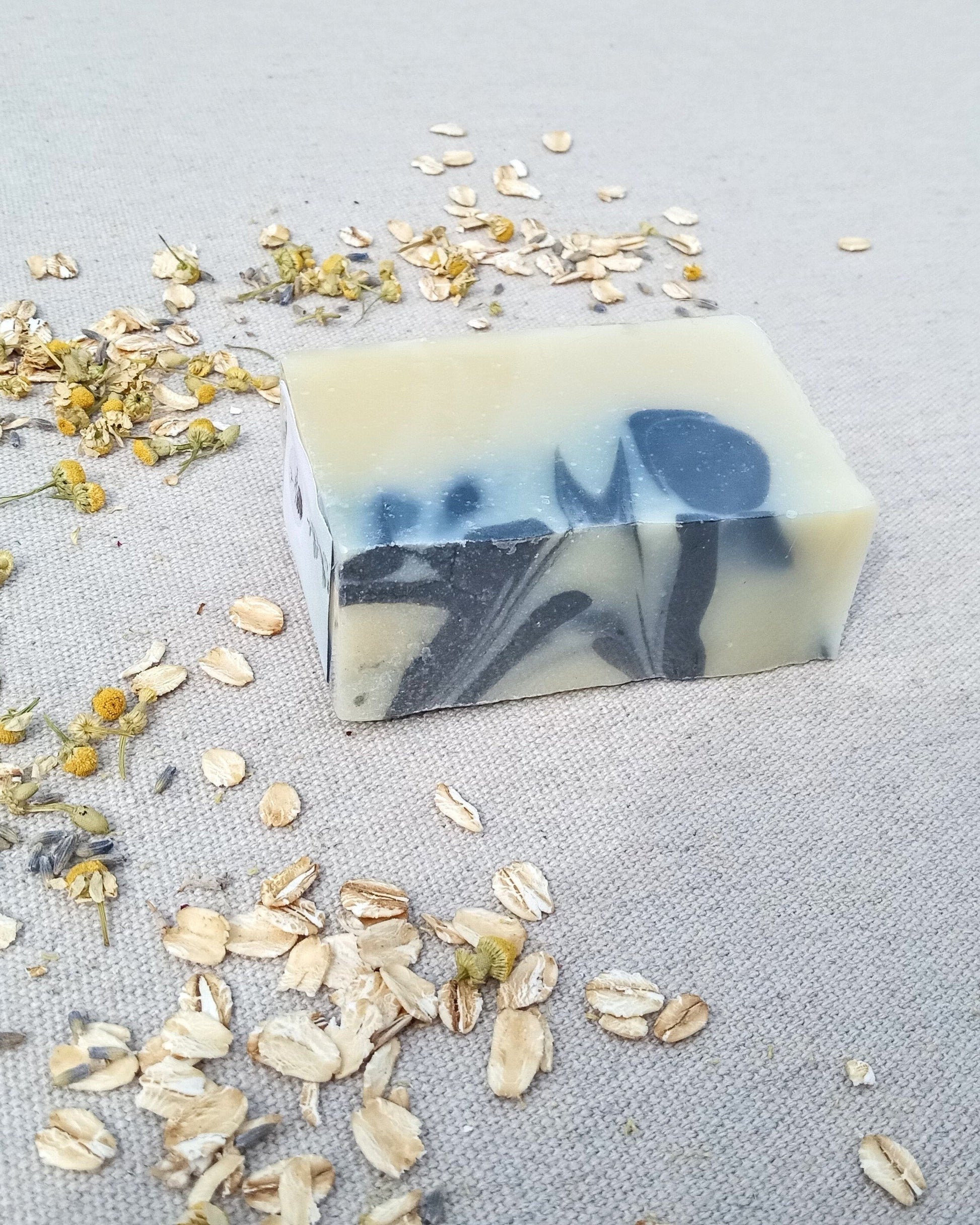 white rectangular bar of soap with swirls of black set on a linen cloth with dried wildflowers and oats scattered around it
