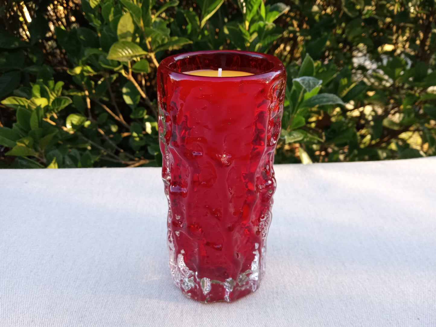 a tall red glass blown cup with a yellow beeswax candle in it wicked with cotton set on a white tablecloth with greenery in the background