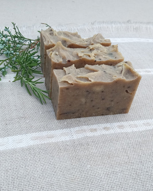 three brown soap bars with flecks of black throughout, rough cut edges and a sculpted top set on linen with a sprig of juniper next to it