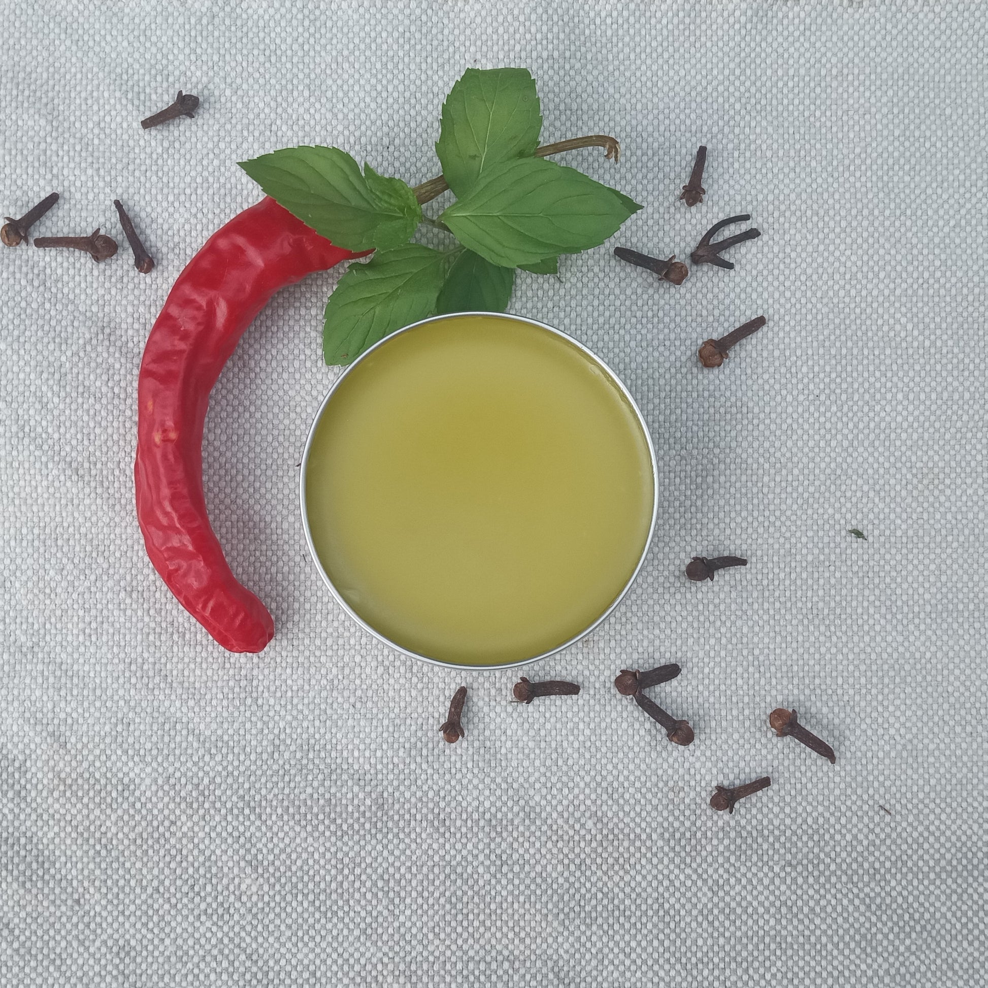 a tin of smooth golden salve set on white linen with a cayenne pepper, cluster of peppermint leaves and dried clove buds scattered around it