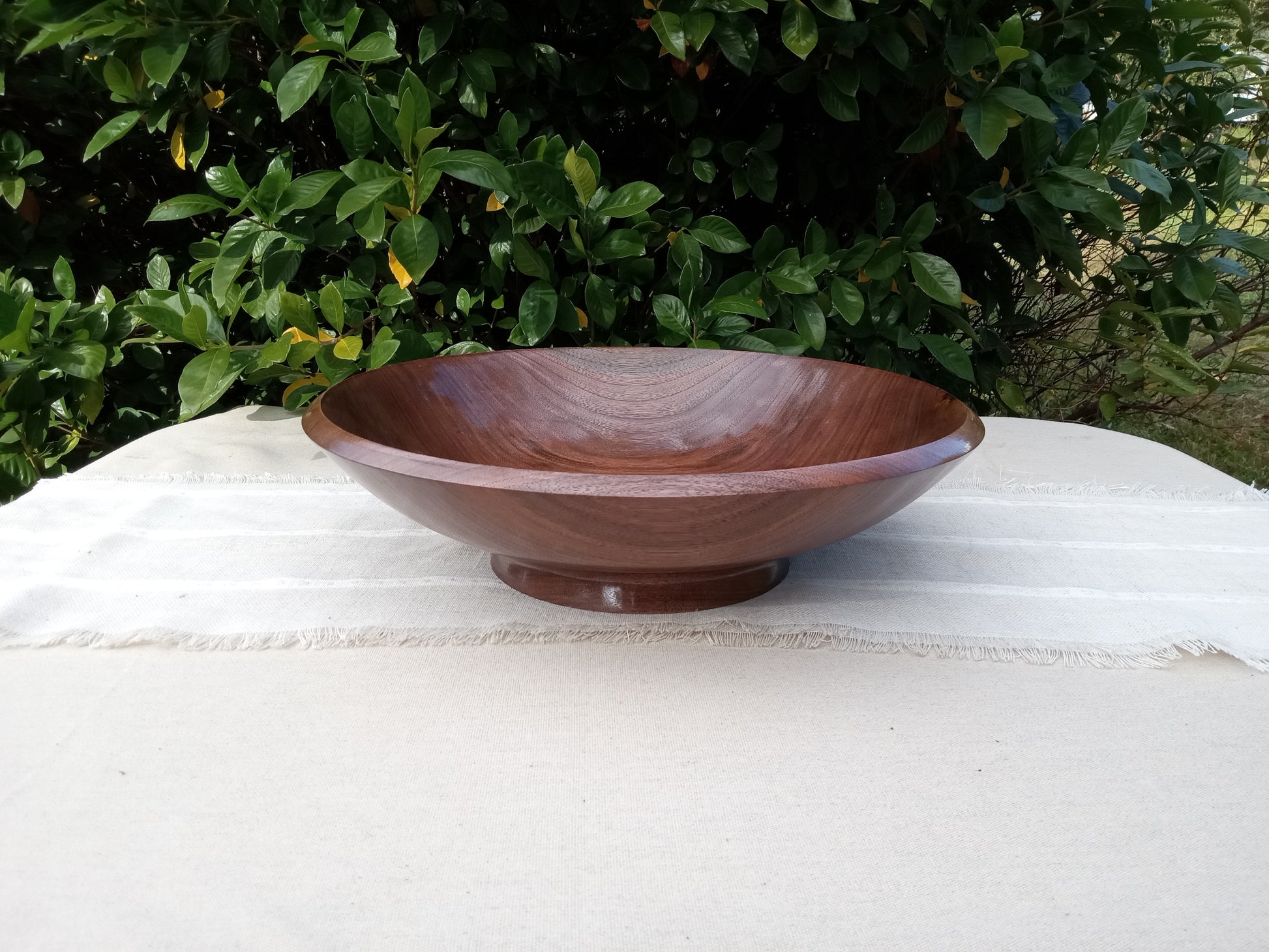 an elegant shallow black walnut wood pedestal bowl with a glossy finish and finely carved rim set on a natural linen table runner with greenery in the background