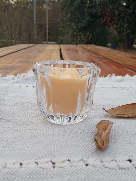 yellow beeswax votive candle in a decorative crystal glass vessel set on a woven table runner atop a rustic wooden table with dried leaves scattered around it