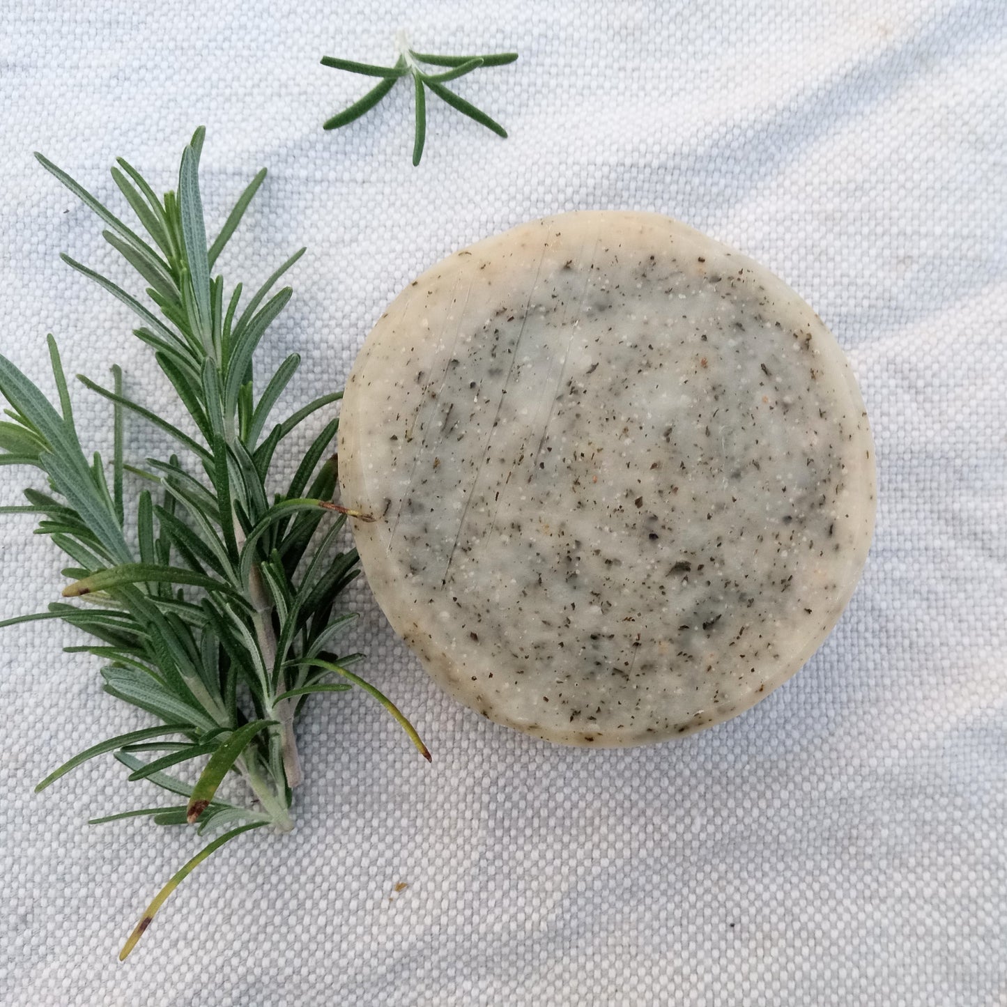 a rustic cream colored round bar of soap with flecks of green scattered throughout it set on a white cloth with sprigs of rosemary beside it
