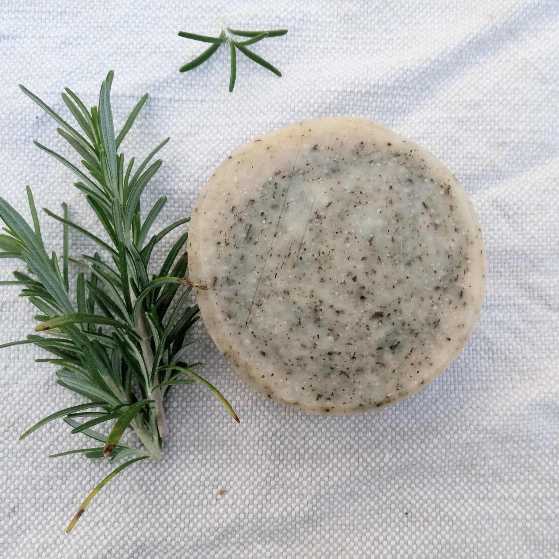 a rustic cream colored round bar of soap with flecks of green scattered throughout it set on a white cloth with sprigs of rosemary beside it