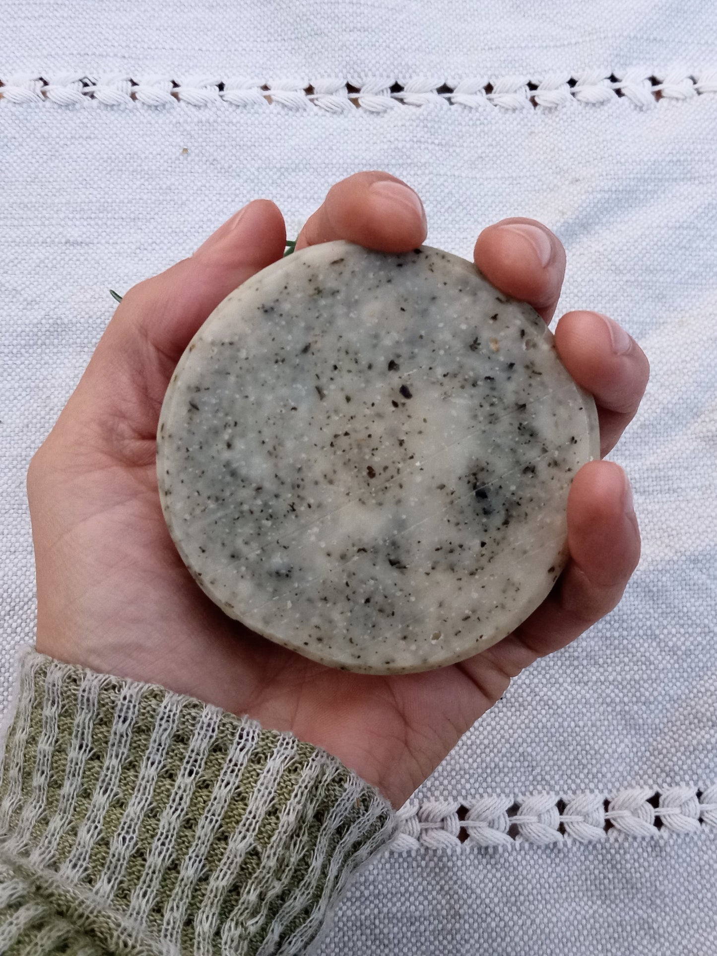 a rustic cream colored round bar of soap with flecks of green in it held in a persons hand with an olive green sweater sleave and a pale blue woven cloth in the background