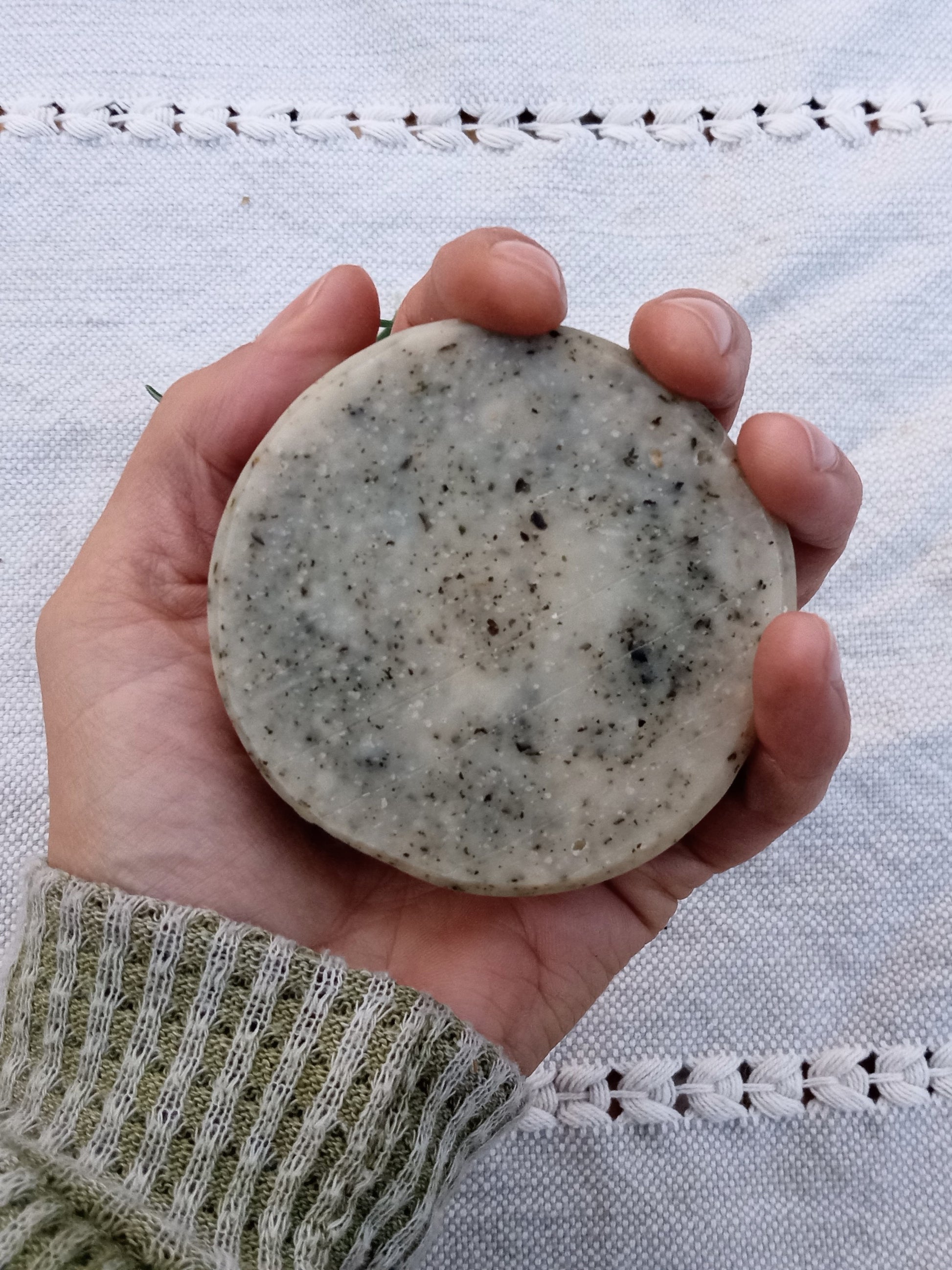 a rustic cream colored round bar of soap with flecks of green in it held in a persons hand with an olive green sweater sleave and a pale blue woven cloth in the background
