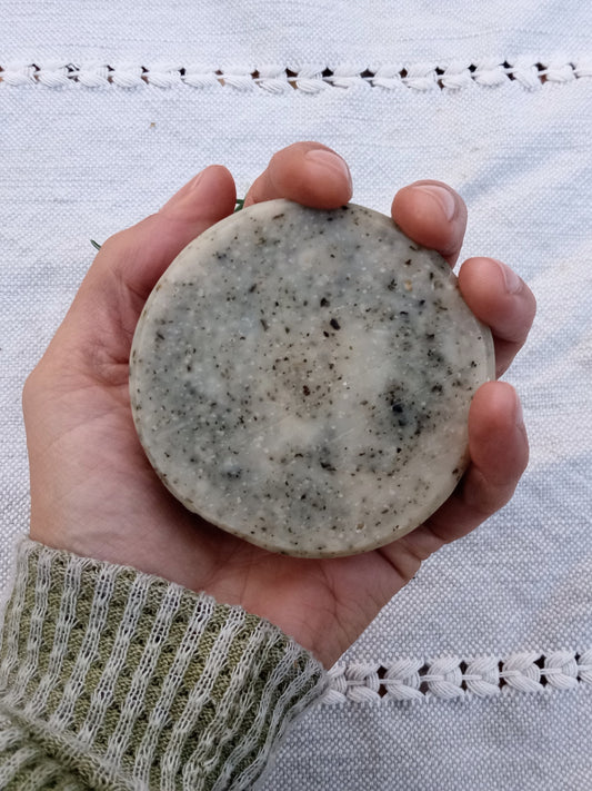 a rustic cream colored round bar of soap with flecks of green in it held in a persons hand with an olive green sweater sleave and a pale blue woven cloth in the background