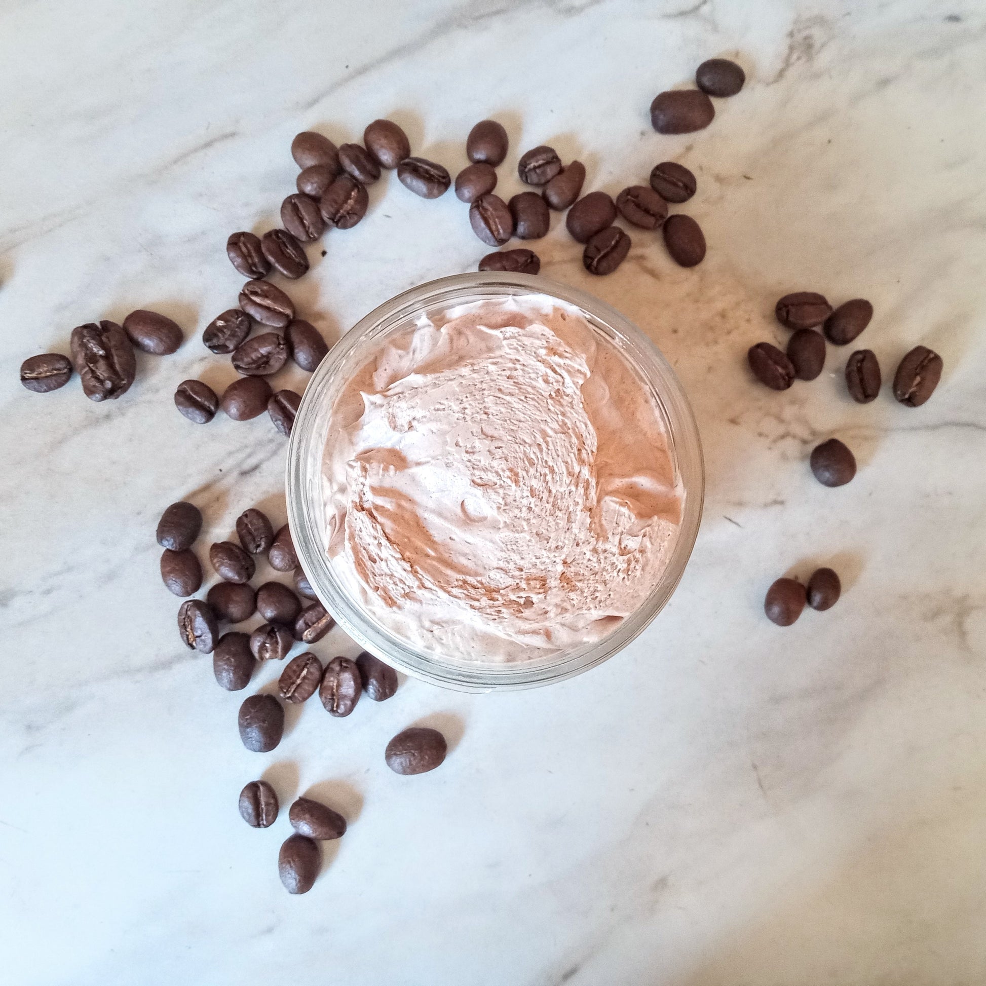 fluffy whipped light brown body butter in a glass jar on a white counter with coffee beans scattered around it