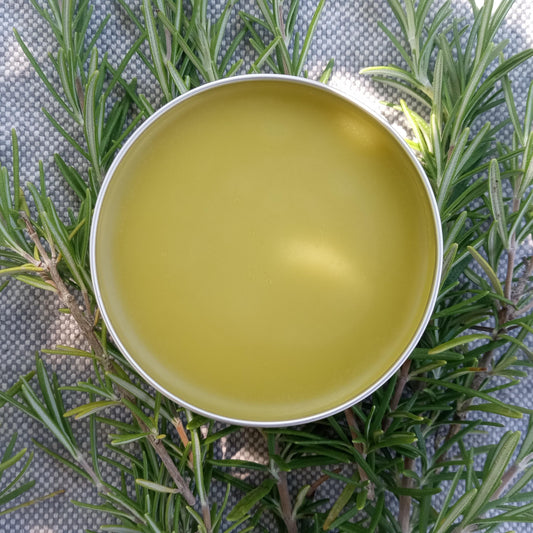pale yellow salve in a silver tin on a pale blue woven fabric with fresh rosemary sprigs around it