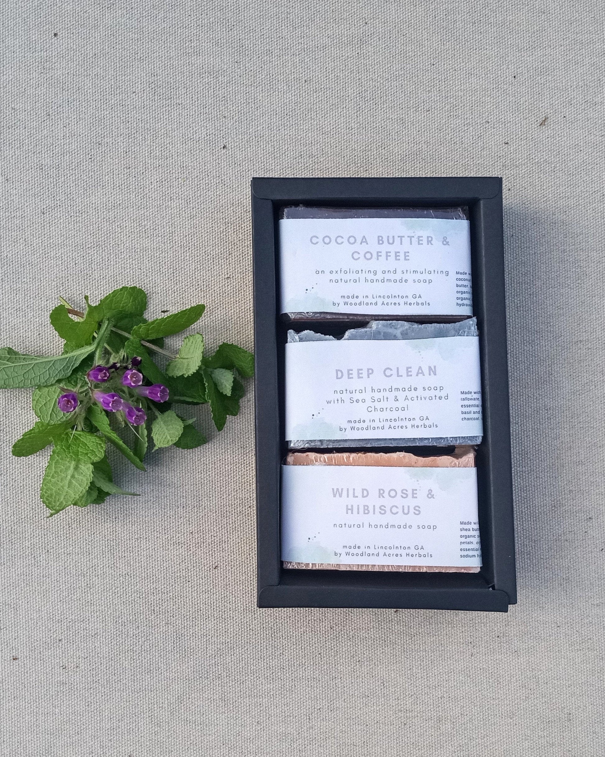 a black drawer box with three wrapped soaps in it set atop a beige cloth with fresh herbs beside it