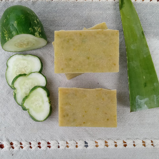 green rectangular soap bars with rustic edges and flecks of deeper green set on a pale blue woven cloth with a sliced cucumber on one side and a fresh aloe leaf on the other