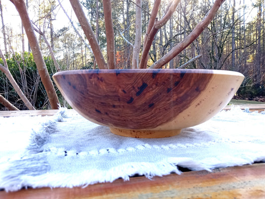 a highly figured hickory burl bowl with deep amber heartwood and cream sapwood contrasts in the sidewall, shaped in an elegant round body design with a pedestal footing set on a woven blue cotton table runner