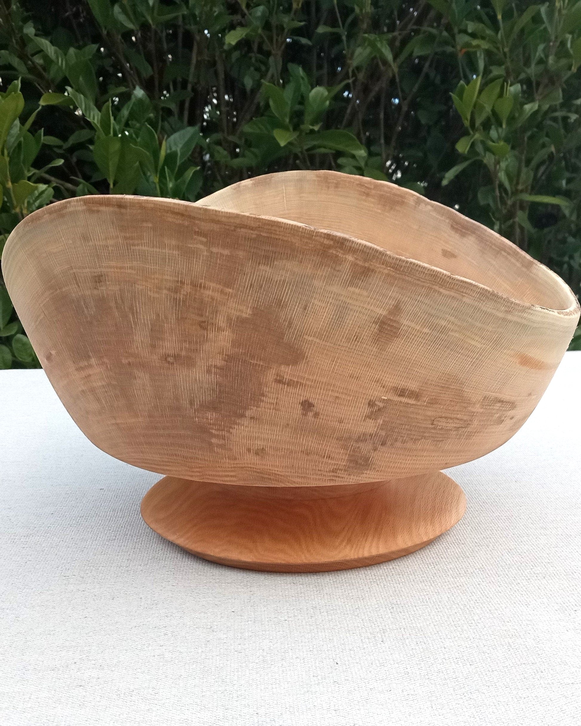 a live edge light wood pedestal bowl with an expertly carved rounded edge base set on a white table cloth with greenery in the background